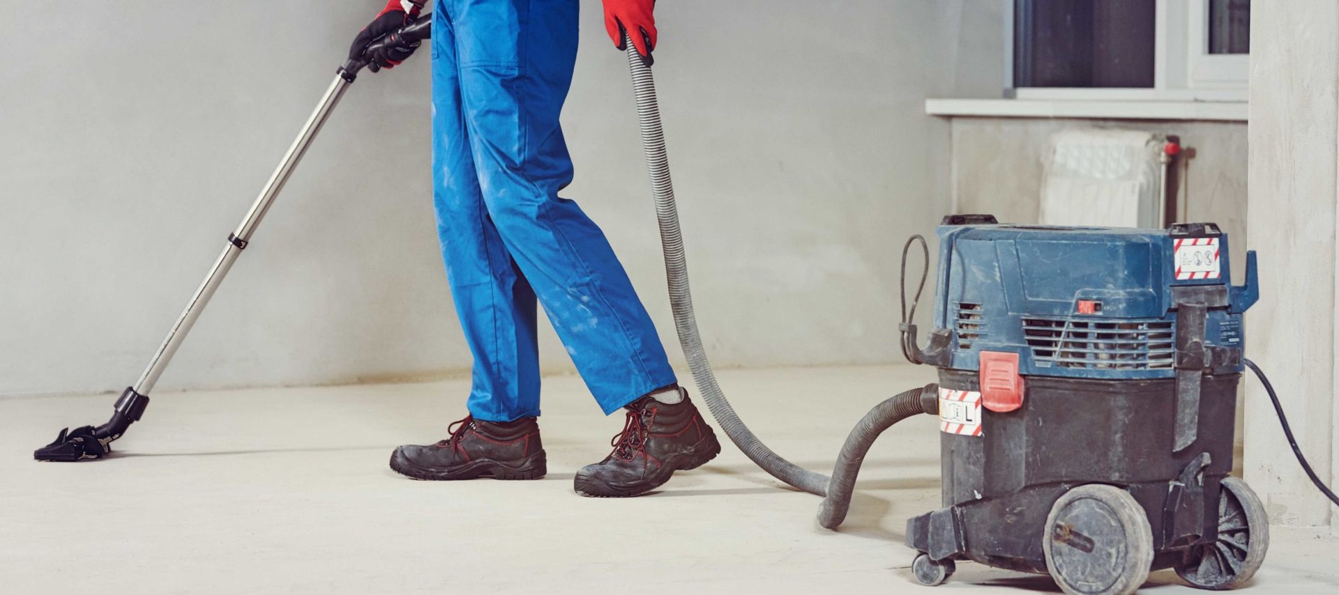 Person in blue overalls vacuuming a light-colored floor with a large shop vac, possibly in a construction setting.