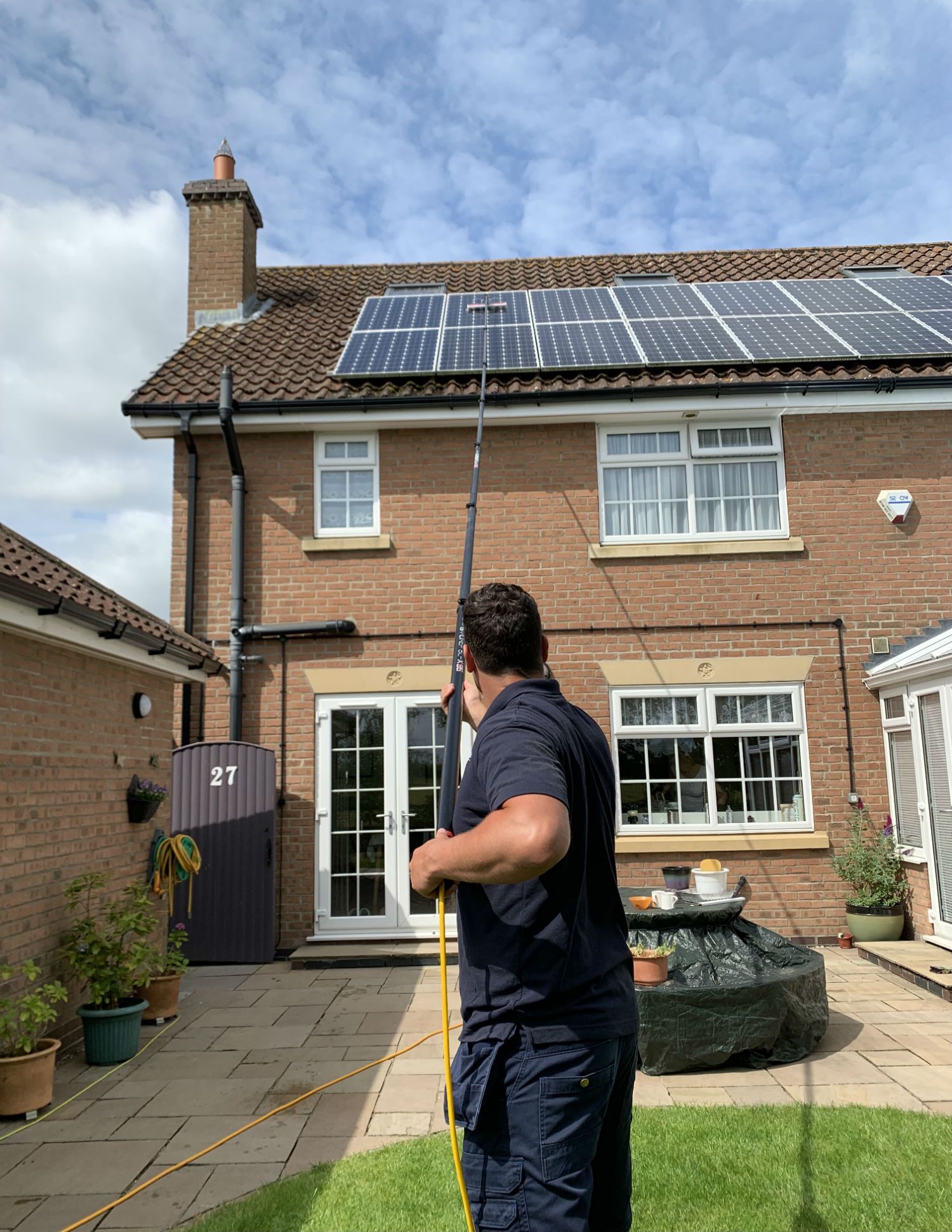 Man cleaning gutters of a two-story brick house with solar panels, using a long pole.