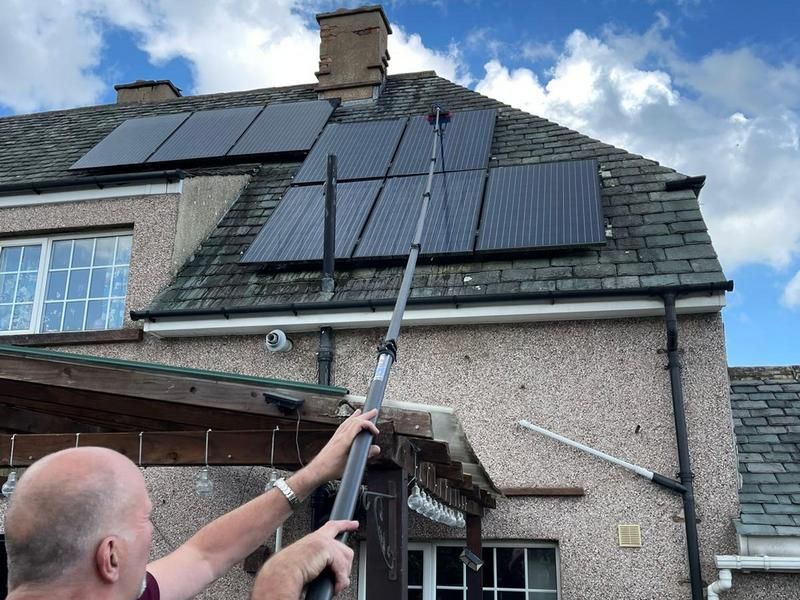 Man cleaning solar panels on a roof with an extendable pole. Cloudy sky background.