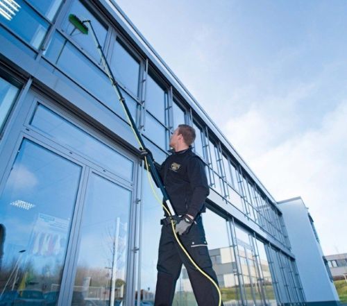 Man cleaning windows of a modern building with a long-handled brush and water hose under a blue sky.