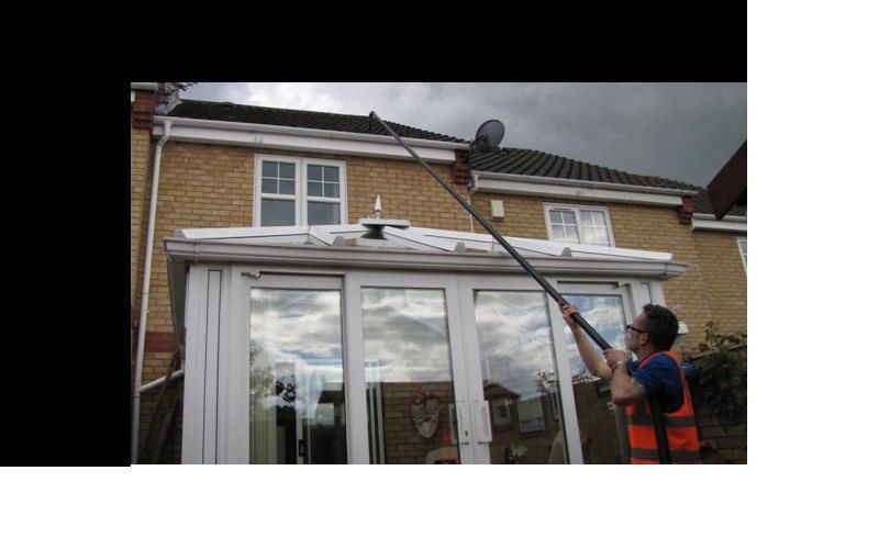 Man cleaning gutters with a long-handled tool on a house with white windows and a conservatory. Overcast sky.