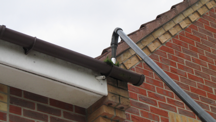 Gutter cleaning tool in use on a brown gutter. The tool is black. Red brick building in the background.