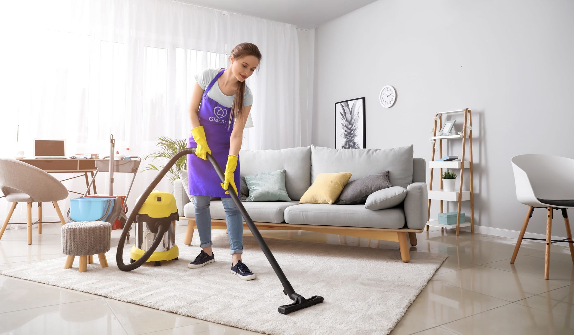 Woman vacuuming a light-colored rug in a bright, modern living room. She wears gloves and an apron, holding the vacuum.