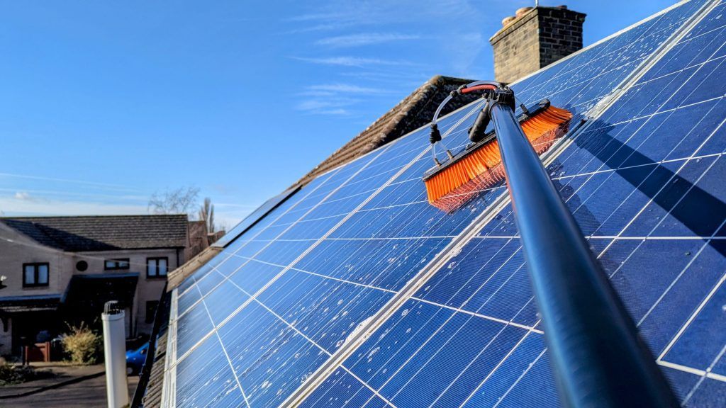 Solar panels being cleaned on a roof with a long-handled brush, blue sky background.