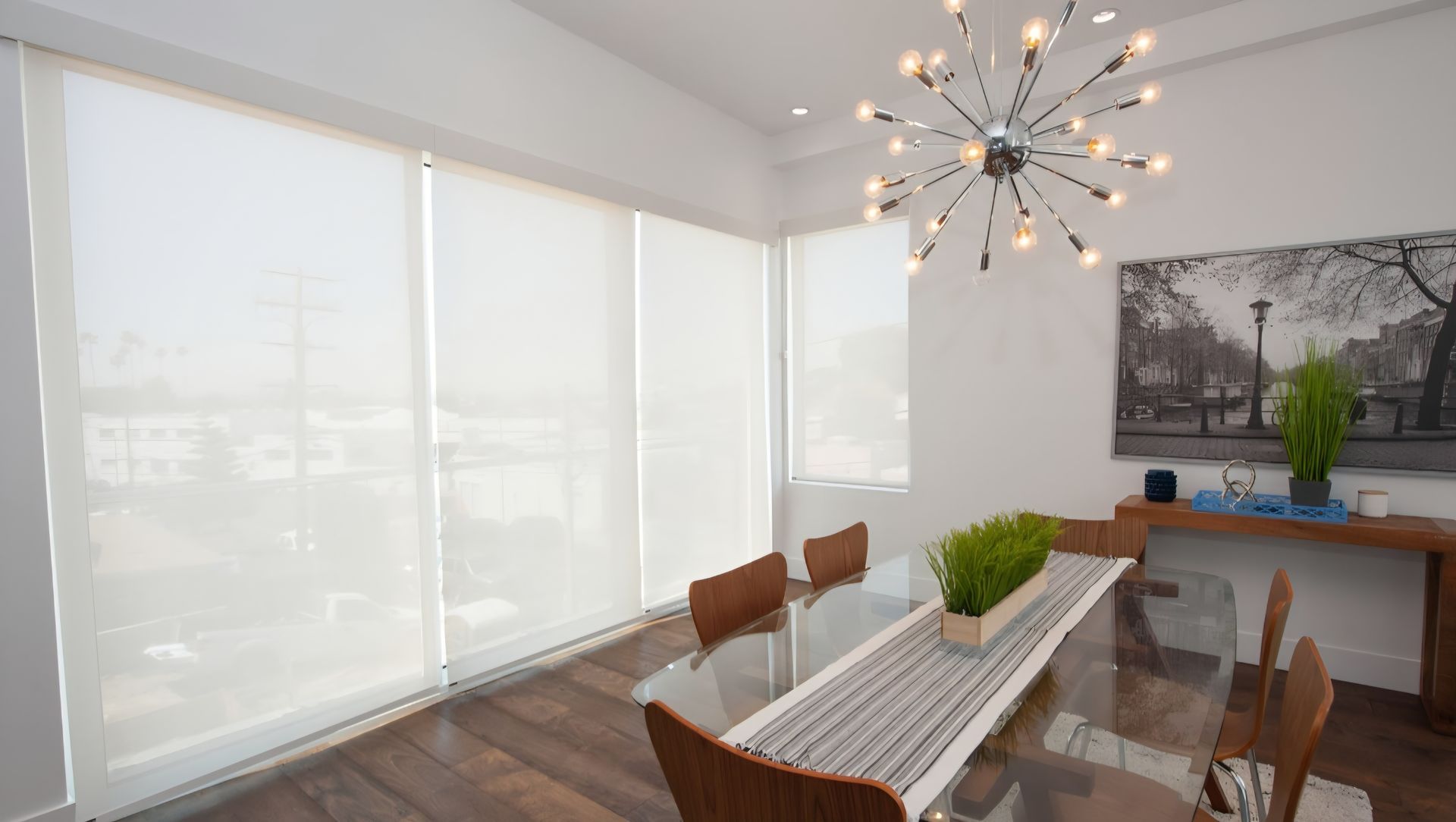 Dining room with white roller shades, glass table, wood chairs, and modern light fixture.