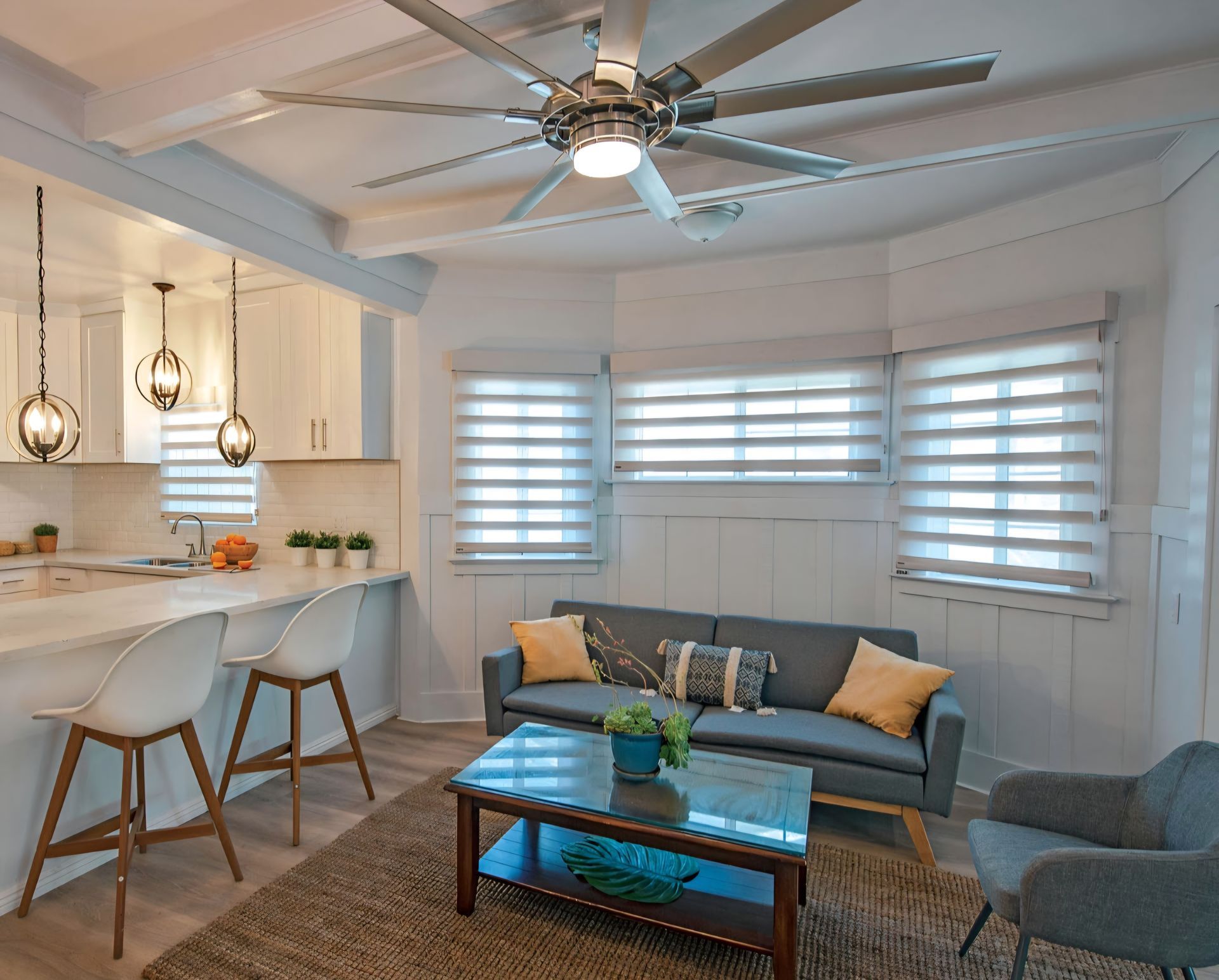 Cozy living room with open kitchen, white walls, gray sofa, coffee table, and large ceiling fan.