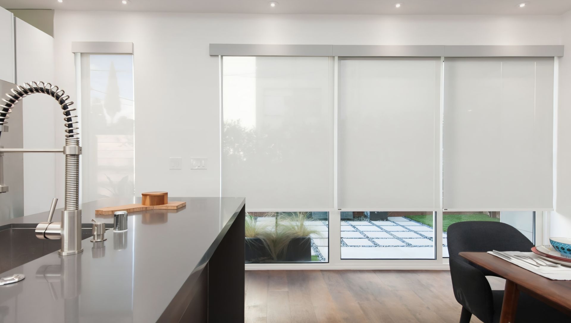 Modern kitchen with white roller blinds, large windows, dark countertop, and dining area.