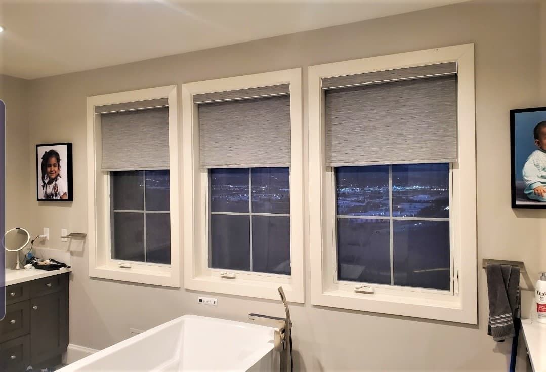 Three windows in a bathroom with roller shades, a bathtub, and pictures on the wall.
