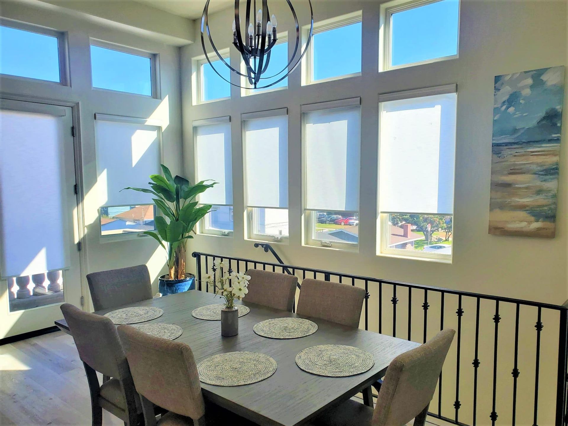 Dining area with a table set for six, large windows with blinds, a plant, and artwork.