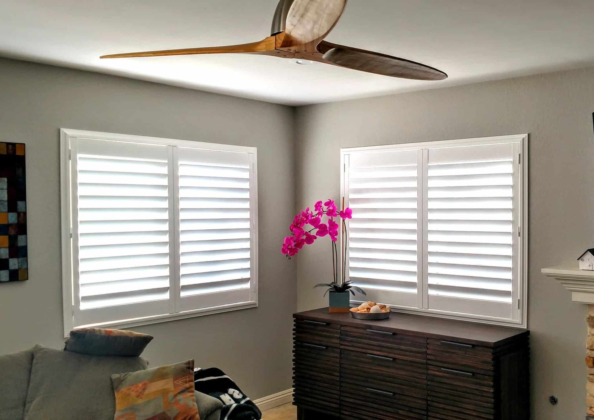White-shuttered windows in a corner room, next to a cabinet with a pink orchid and a ceiling fan.