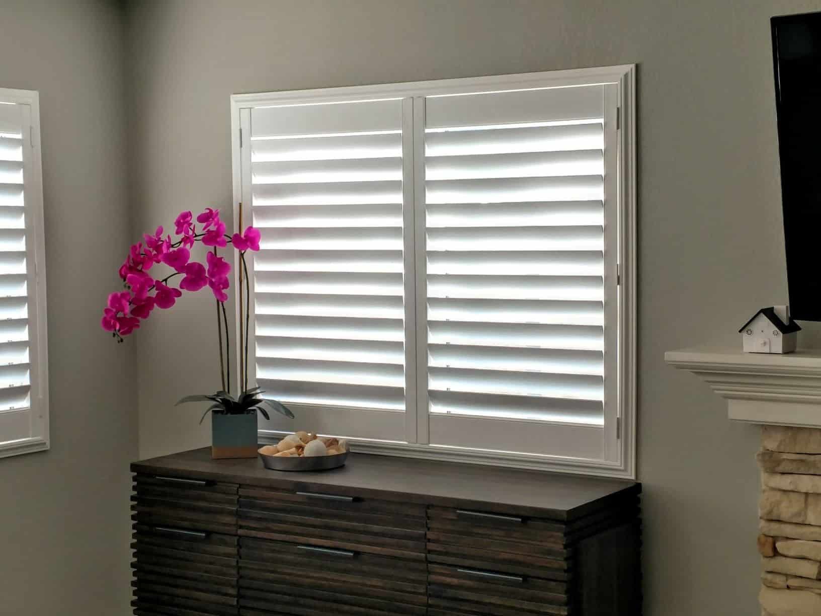 White shutters on a window above a dark wood cabinet with a pink orchid.