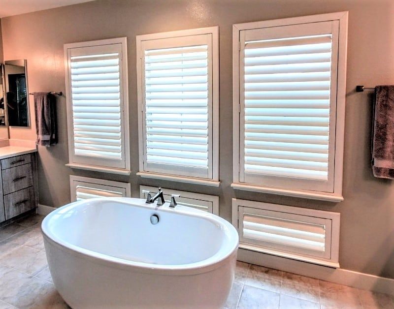 White bathtub in front of three windows with white shutters. Bathroom with gray walls and towels.