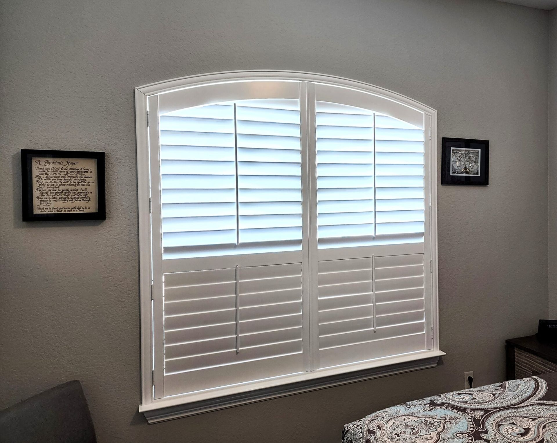 White shutters on an arched window in a room with gray walls, two framed pictures, and a patterned bed.
