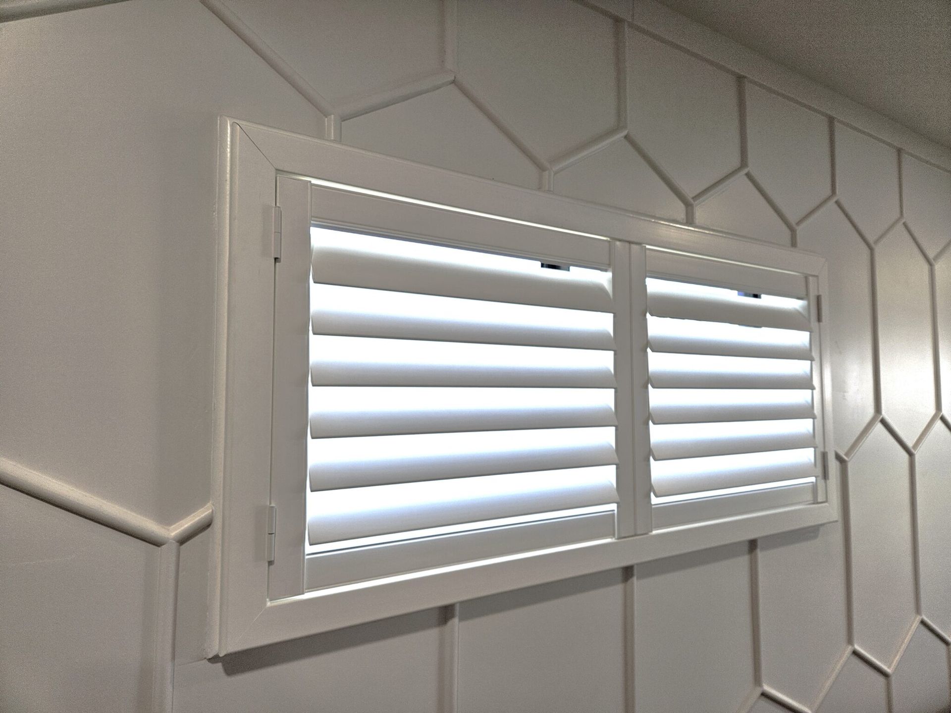 White shutters over a window on a wall with white geometric paneling.