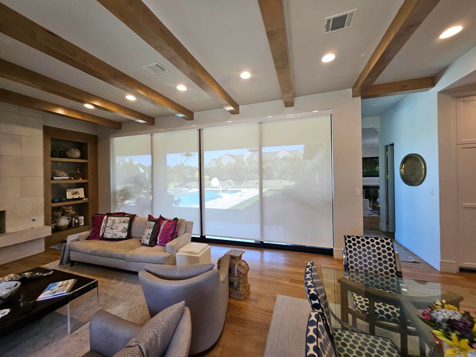 Living room with large windows, wood beams, and neutral furnishings overlooking a pool.