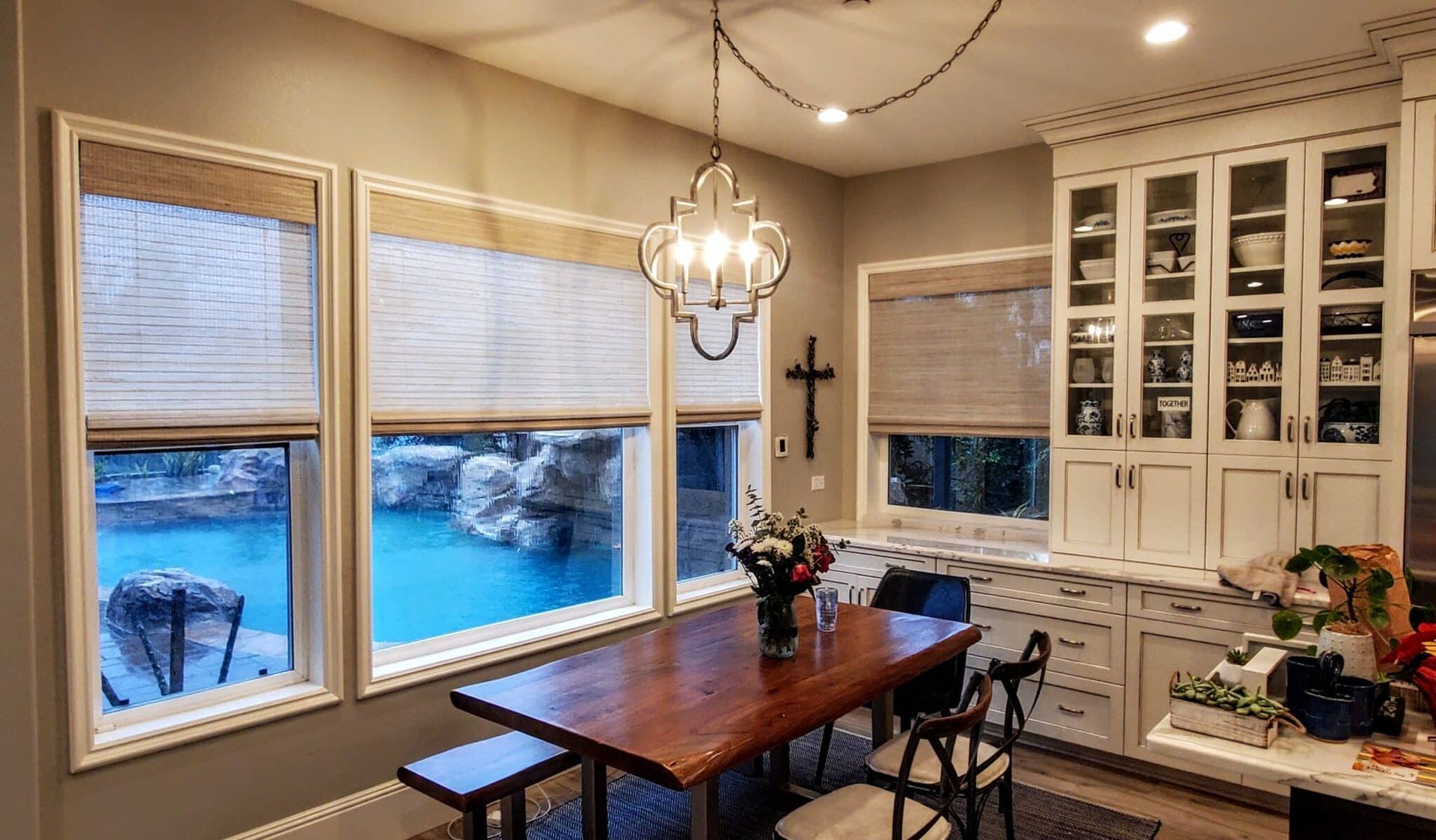 Dining room with table by windows overlooking a pool. Kitchen cabinets and chandelier.