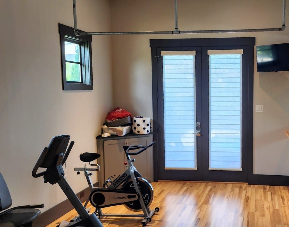 A home gym with a stationary bike, doors with shades, a small window, and wooden floors.