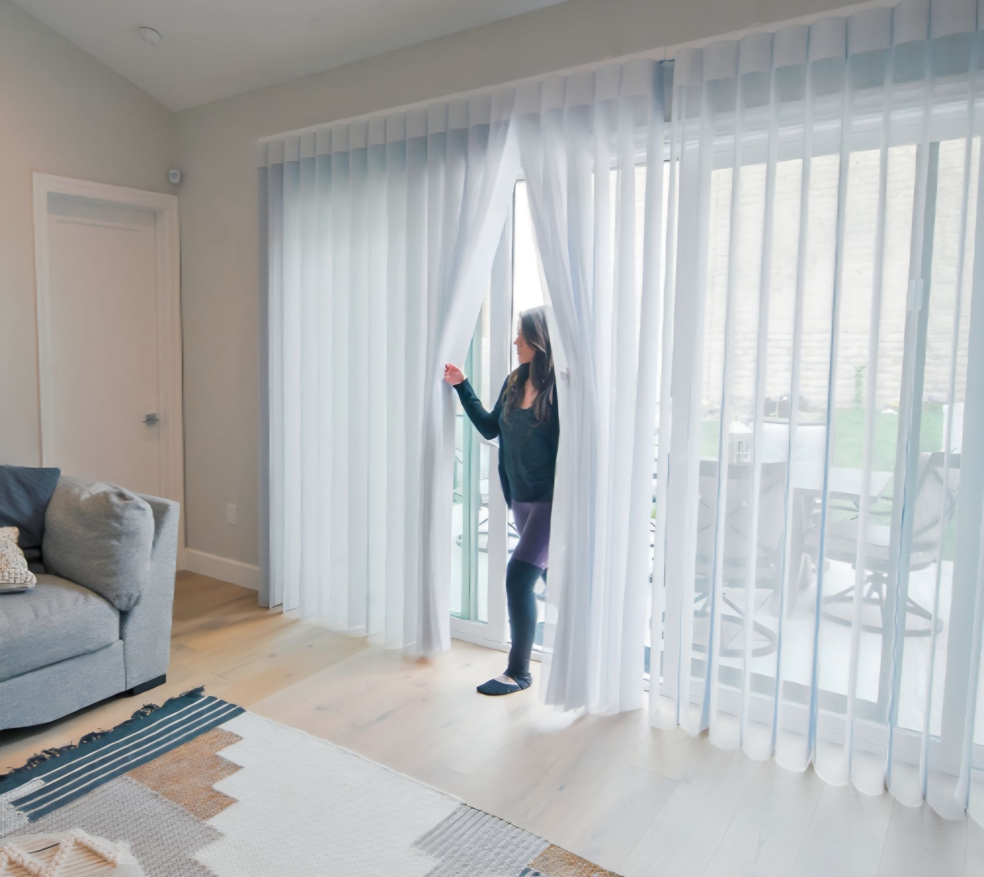Woman opens sheer white curtains on a sliding glass door, revealing a patio. Interior view.