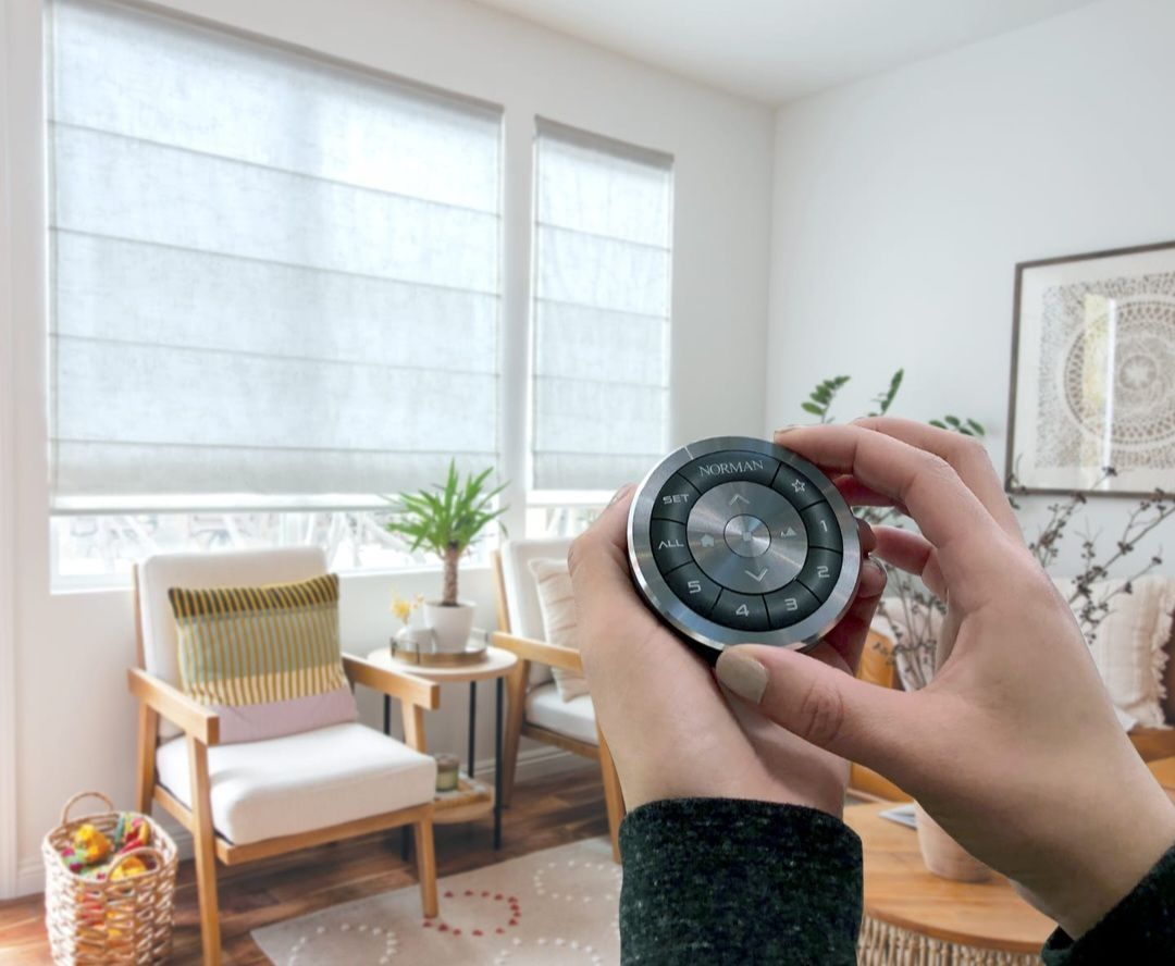 Hands holding a silver smart home device, controlling blinds in a bright living room.