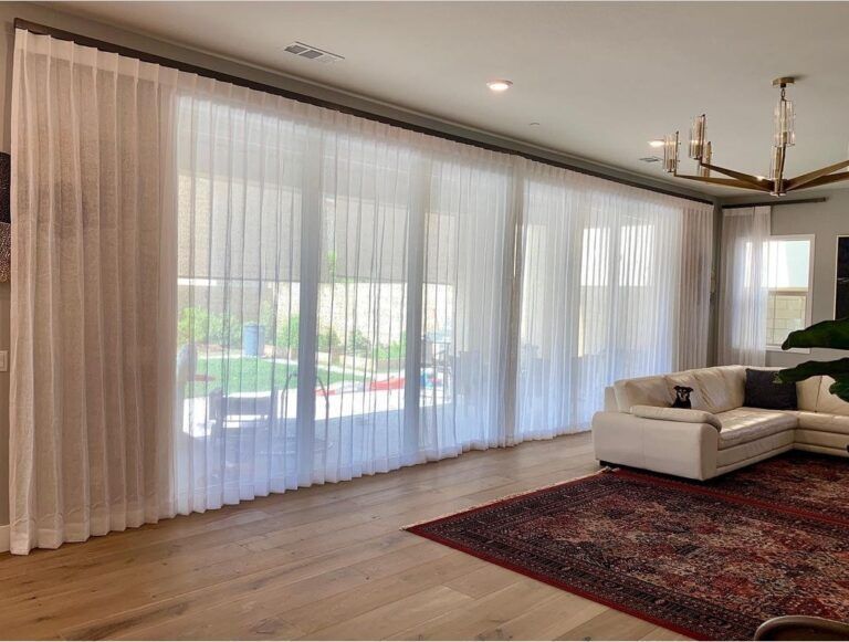 Sheer white curtains covering large windows in a bright living room, over a wood floor, with a white sofa and a red rug.