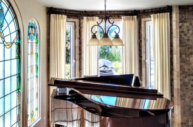 Grand piano in a bright room with a bay window and stained glass, cream curtains, and a chandelier.