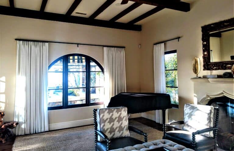 Living room with arched window, white drapes, dark-trimmed windows, black piano, and armchairs with patterned cushions.