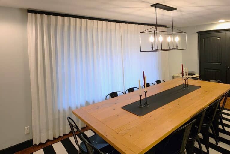 Dining room with a long wooden table, black chairs, and white curtains. A black and white striped rug is below.