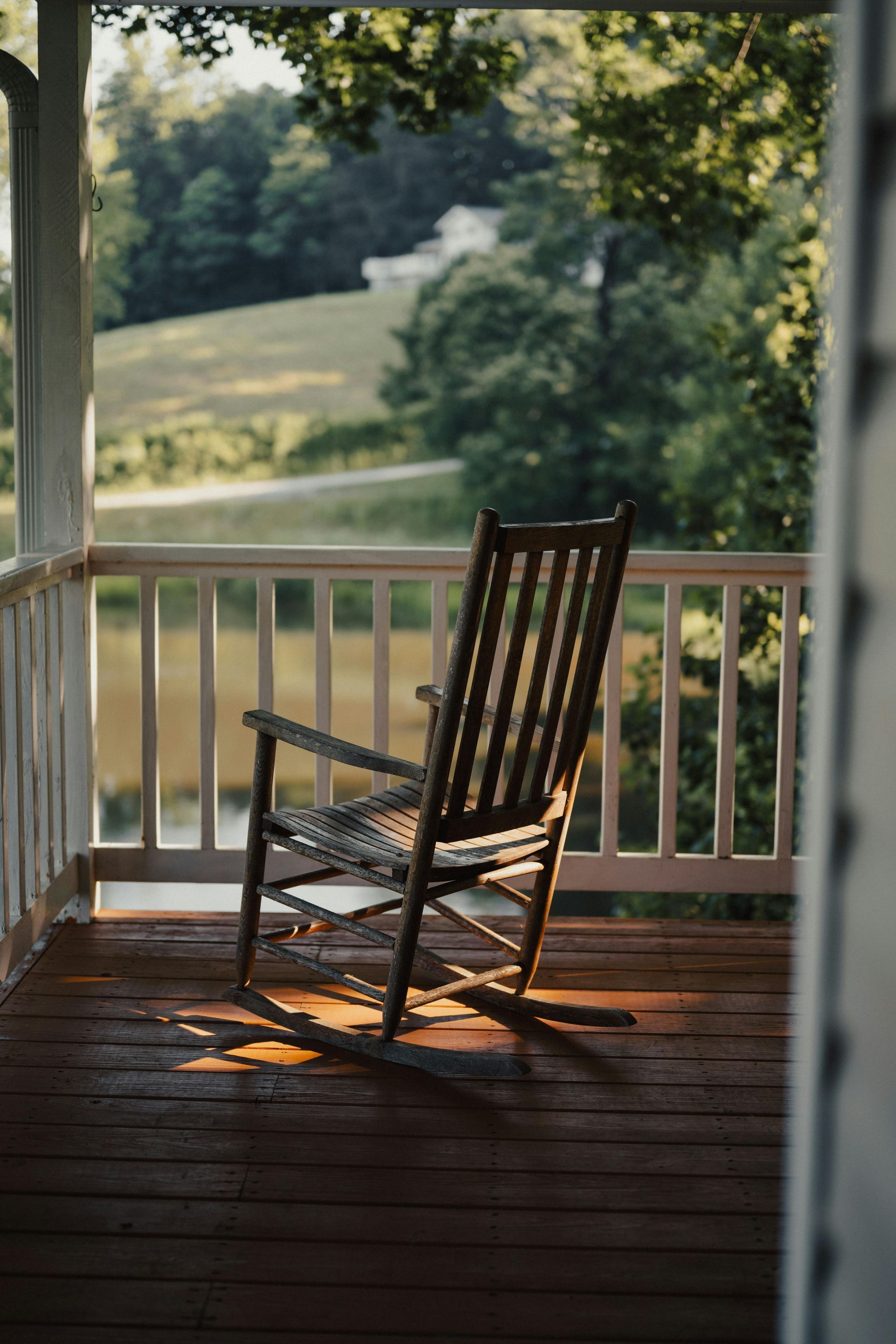 Wooden rocking chair on porch, overlooking a green landscape. Warm sunlight illuminates the scene.