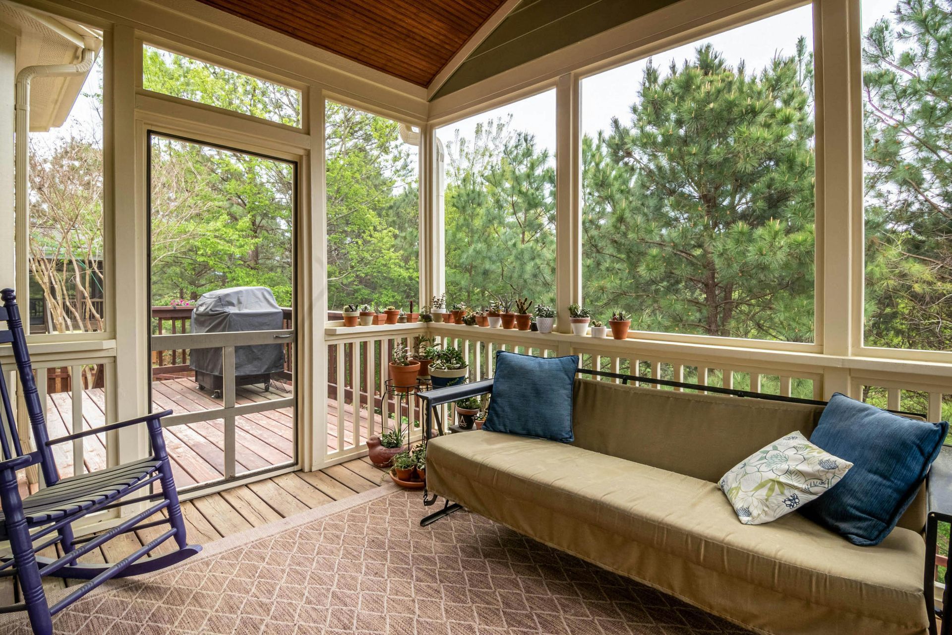 Screened-in porch with seating, plants, and a view of trees. Features rocking chair and outdoor grill.