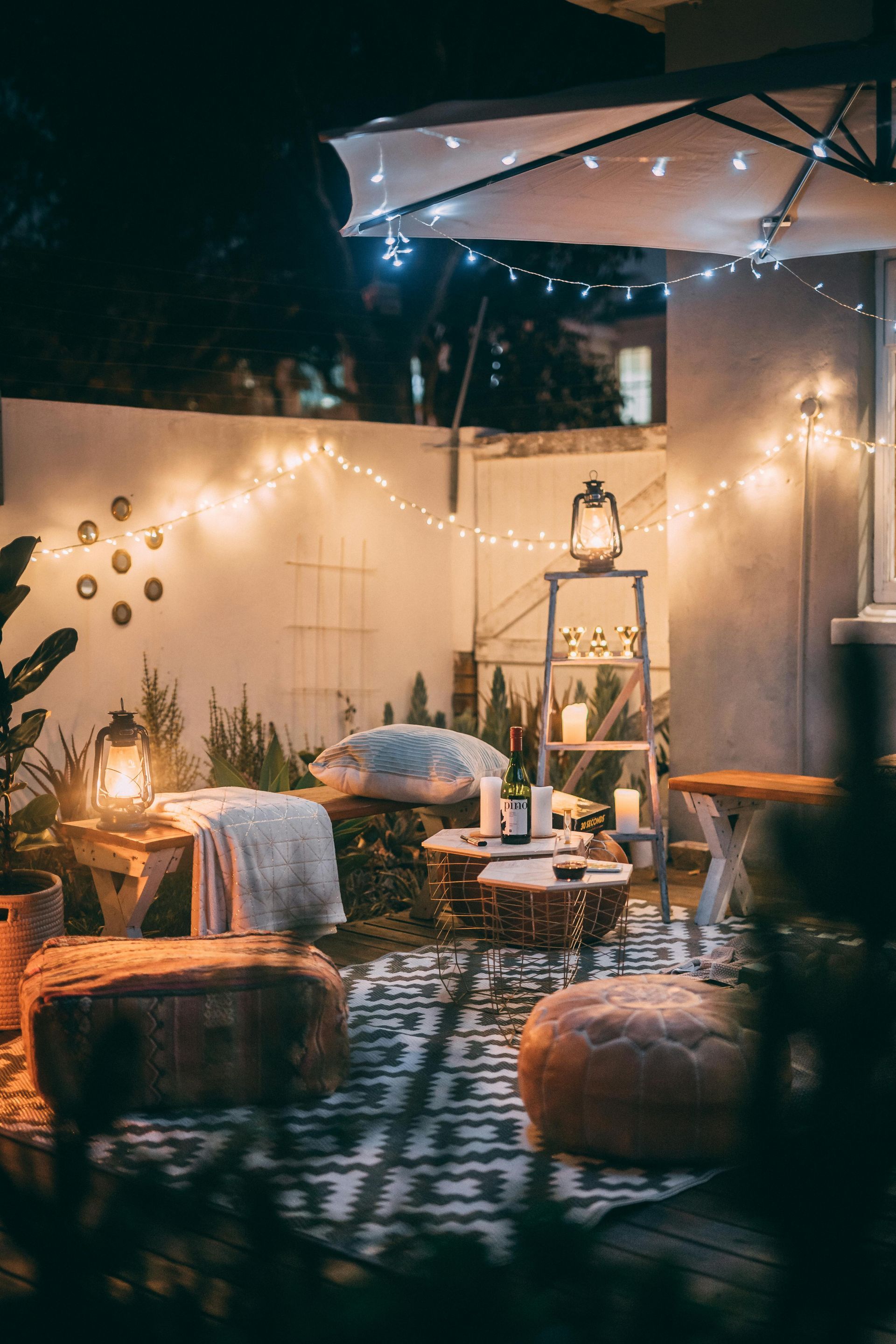 Cozy outdoor patio at night, string lights, lanterns, pillows, rug, neutral colors.