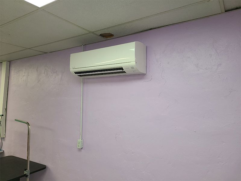 Wall-mounted white air conditioning unit on a purple wall in a room, with electrical cord hanging down.