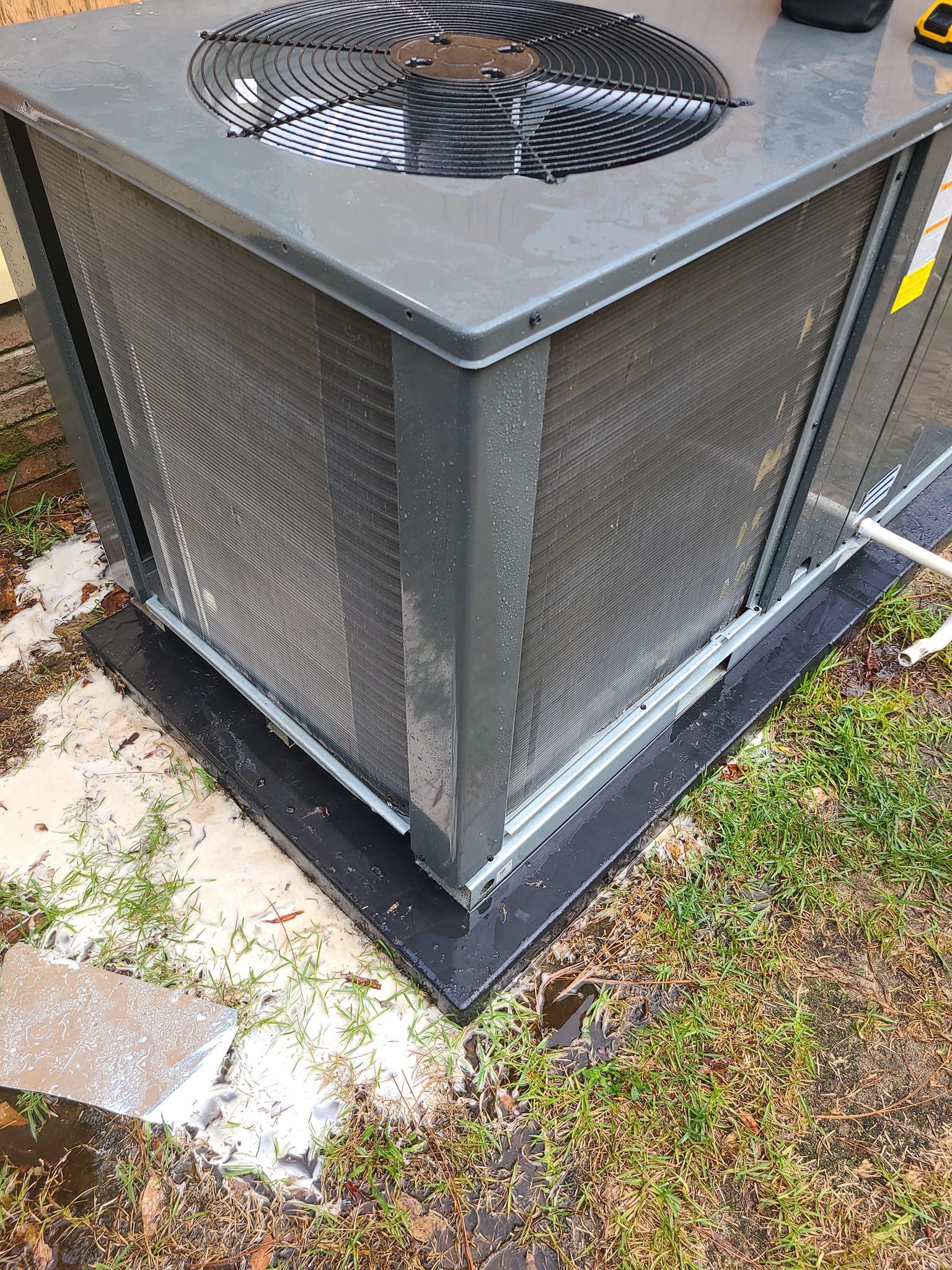 Air conditioner unit with fan on top, metallic gray, set on a black pad outside.