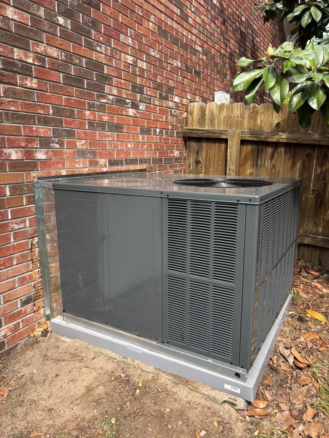 Gray HVAC unit next to a brick wall and a wooden fence, set on a concrete platform.