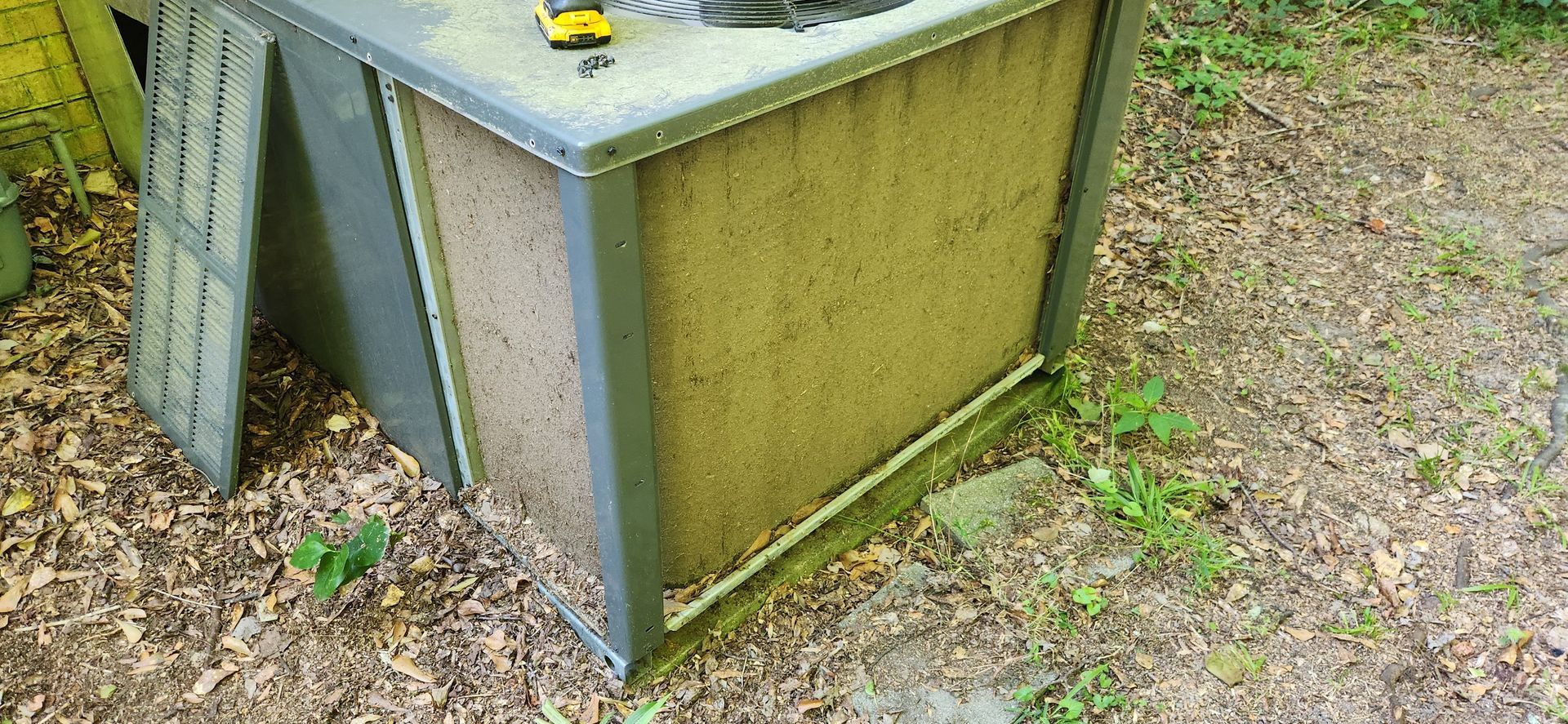 A weathered, open-sided outdoor air conditioner unit, set in a yard with leaves.