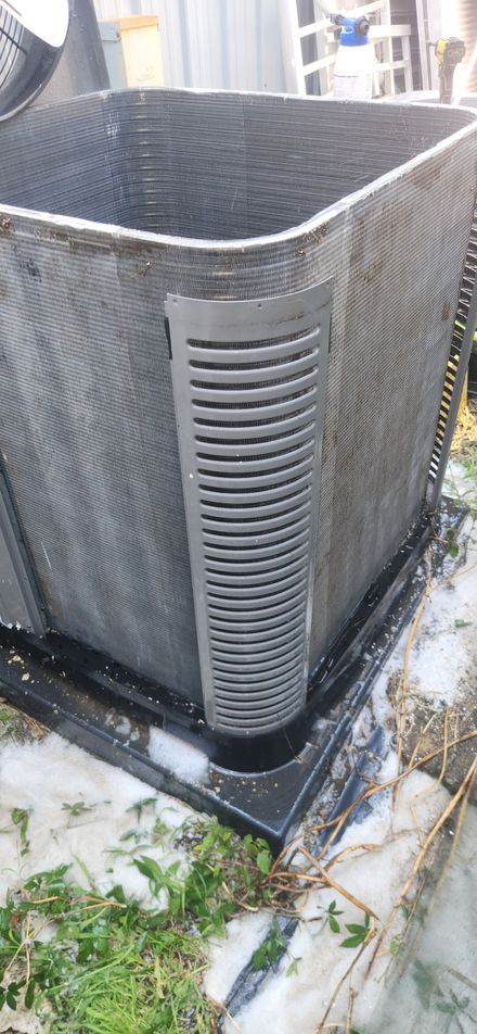 A close-up of a black rectangular air conditioning unit with a vertical vent on its side. White snow surrounds the unit.