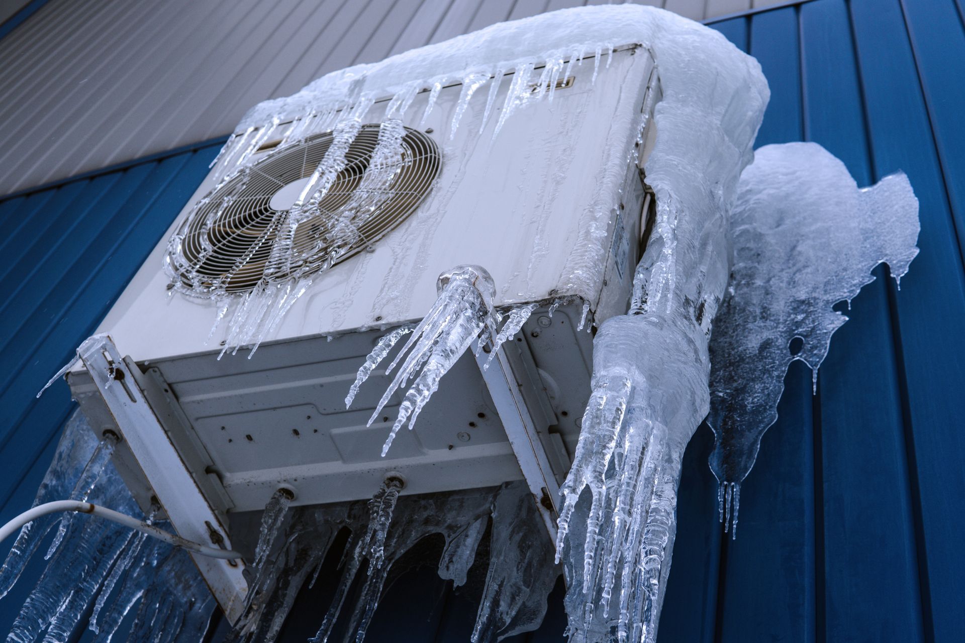 Iced-over air conditioner unit mounted on a blue building wall, covered in frozen icicles.
