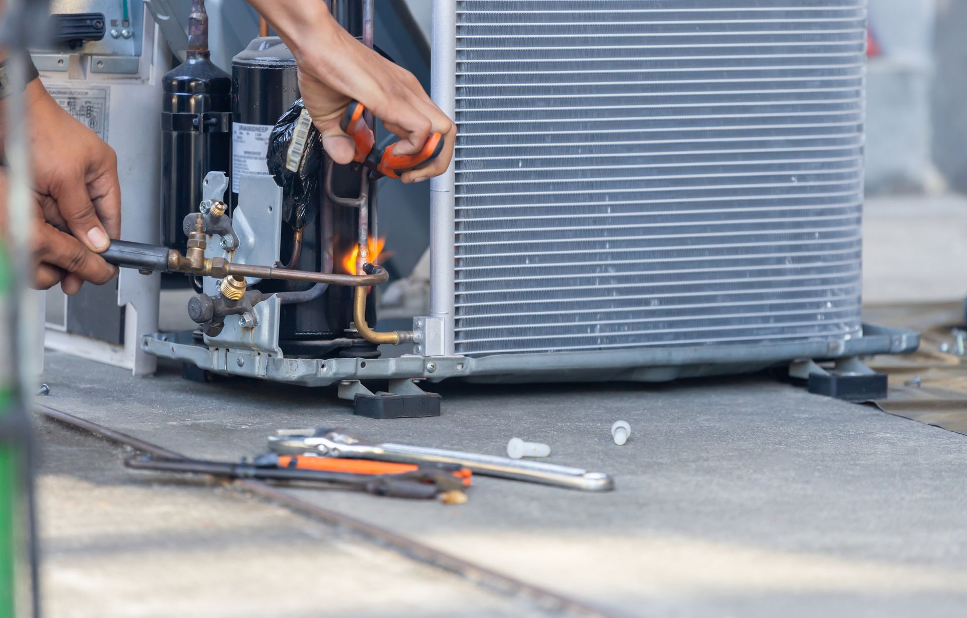 Worker using a torch near an outdoor HVAC unit, with tools on the ground.