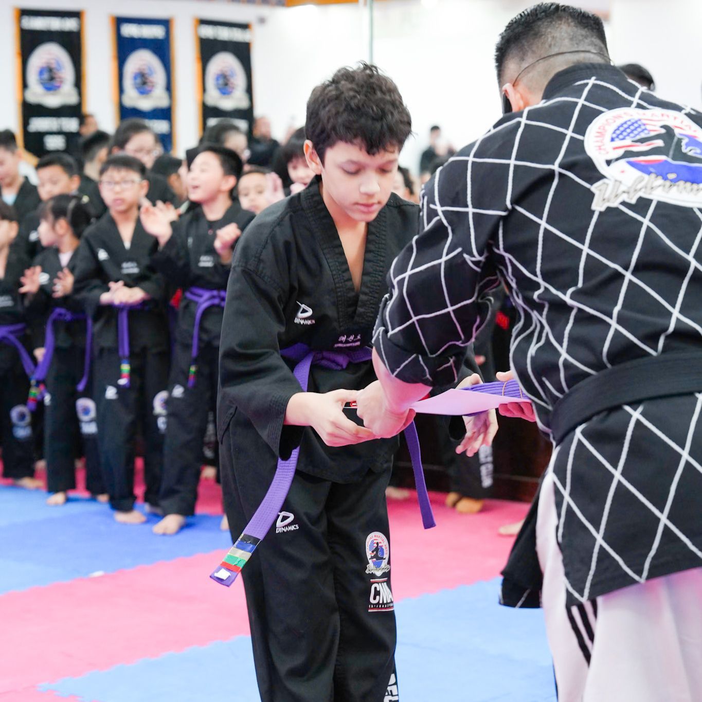 A man in a black and white checkered shirt is giving a boy a purple belt