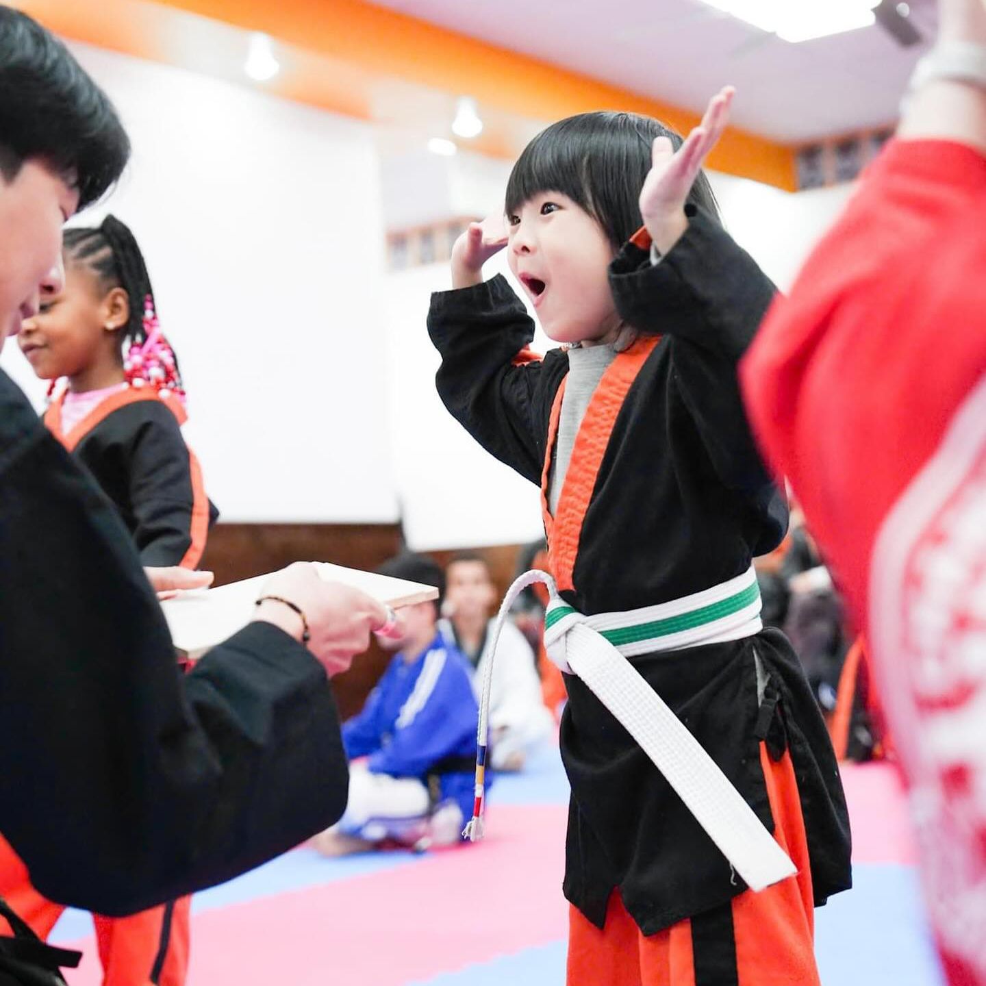 A little girl in a black and orange karate uniform is being taught by a man