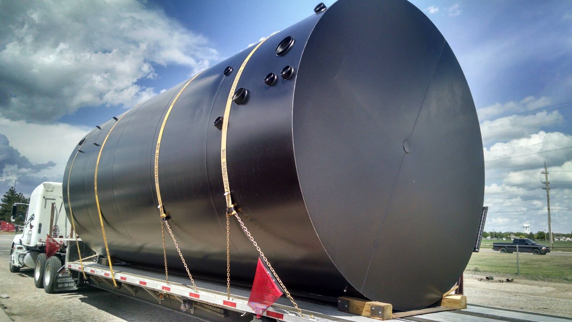 Black cylindrical tank secured on a flatbed truck for transport, under a cloudy sky.