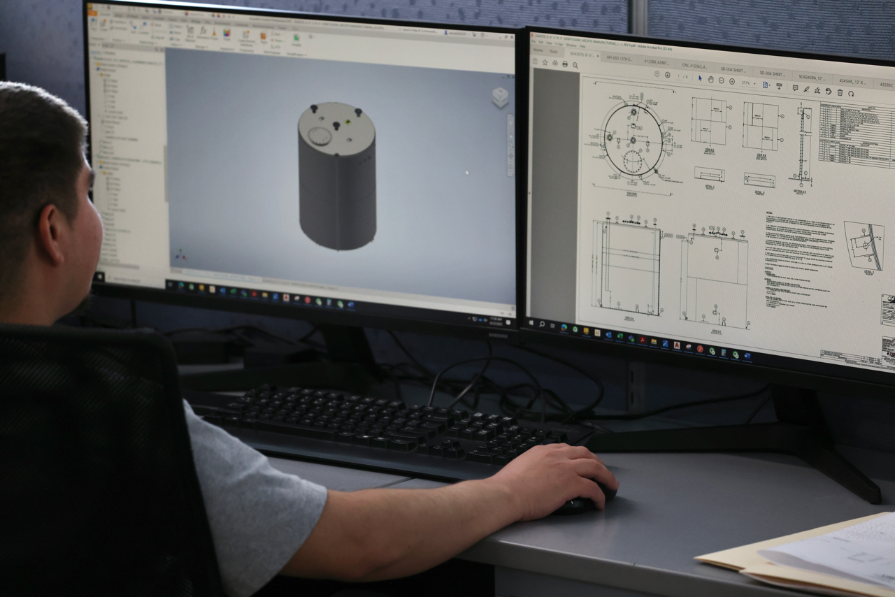 Person working on a computer with dual monitors; one shows 3D model, the other blueprints.