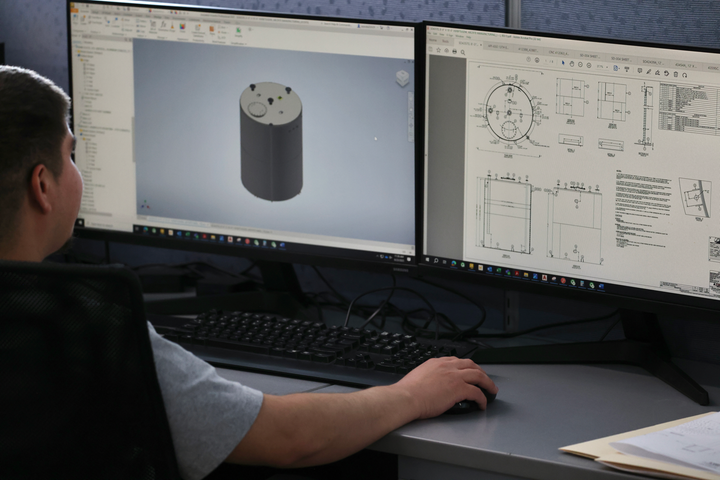 Person working on a computer with dual monitors; one shows 3D model, the other blueprints.