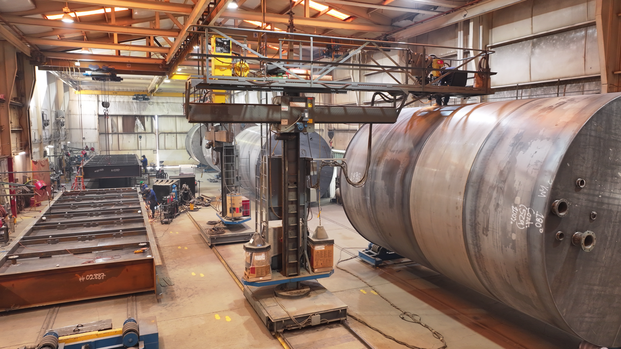 Industrial workshop with large cylindrical metal tanks. Overhead crane, welding equipment, and scaffolding are visible.