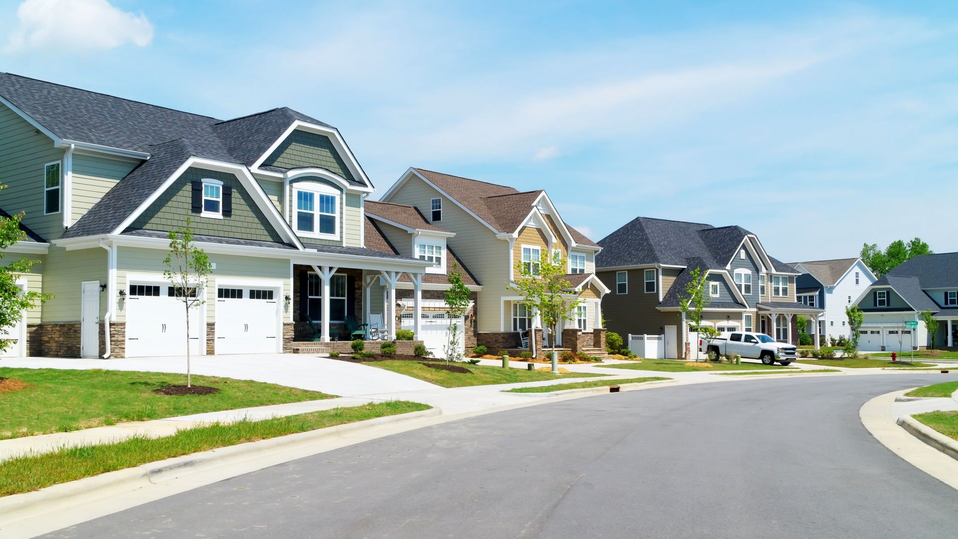 A row of suburban houses with driveways, green lawns, and trees along a paved street under a blue sky.