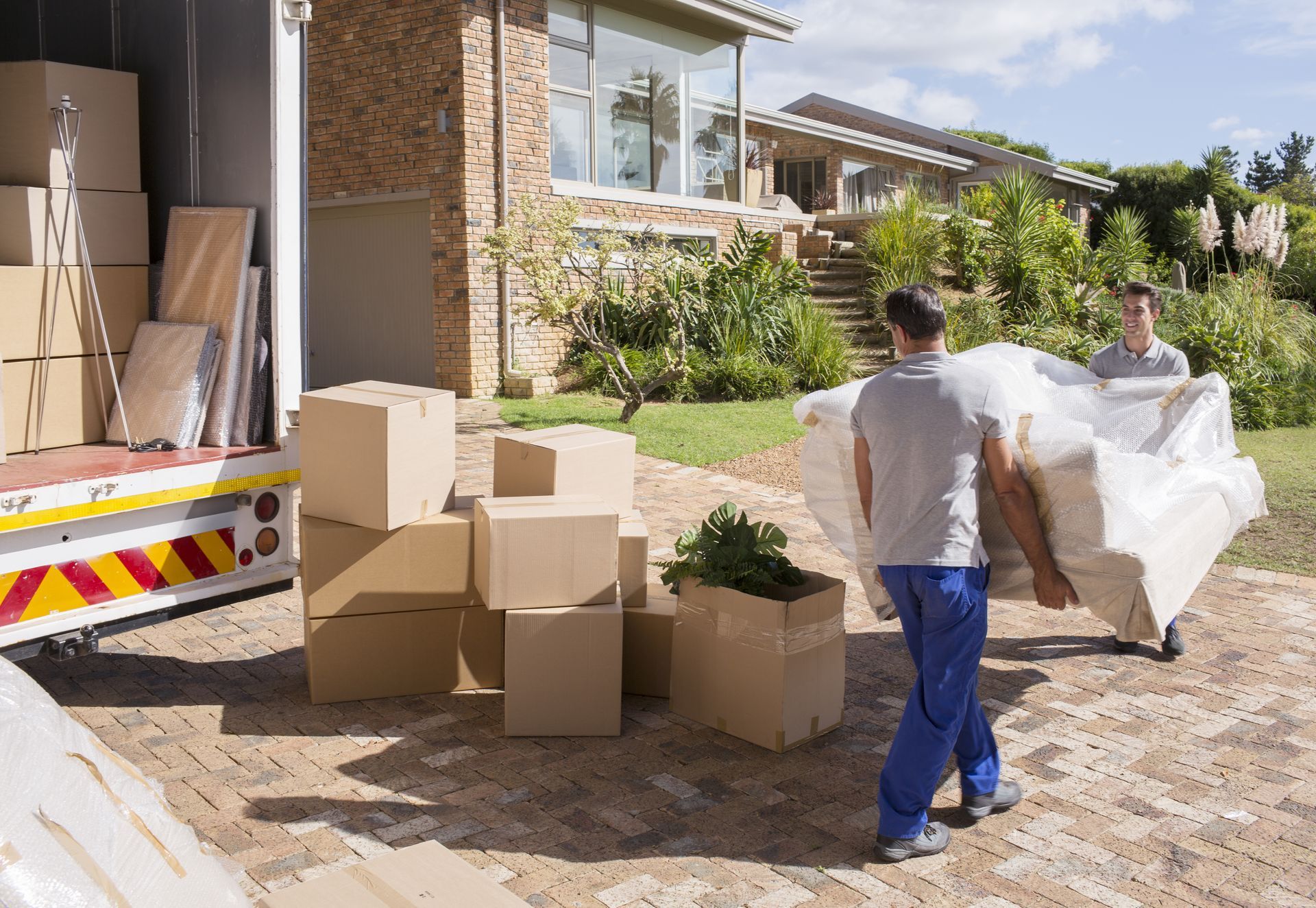 Two movers carry a white-wrapped sofa outside a house with moving boxes stacked near a delivery truck.