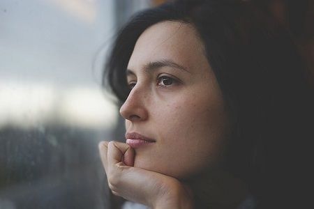 A woman is looking out of a window with her hand on her chin.