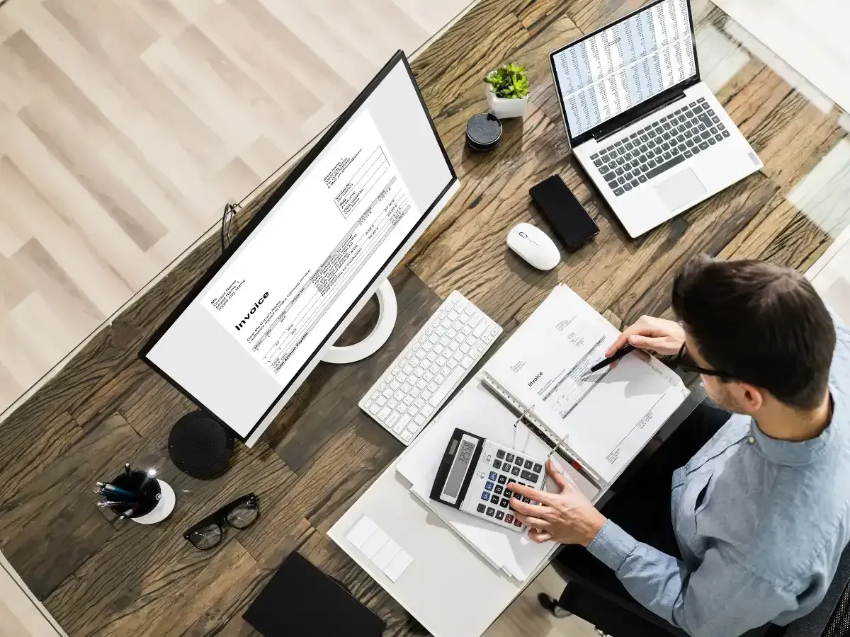 Person reviewing invoices and calculations at a desk while handling insurance bookkeeping tasks on a computer.