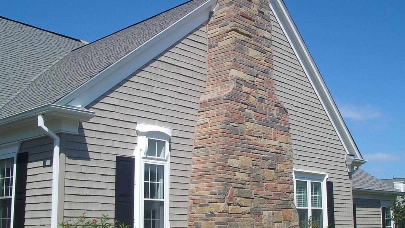 A stone chimney on the side of a gray-sided house with white trim against a bright blue sky.