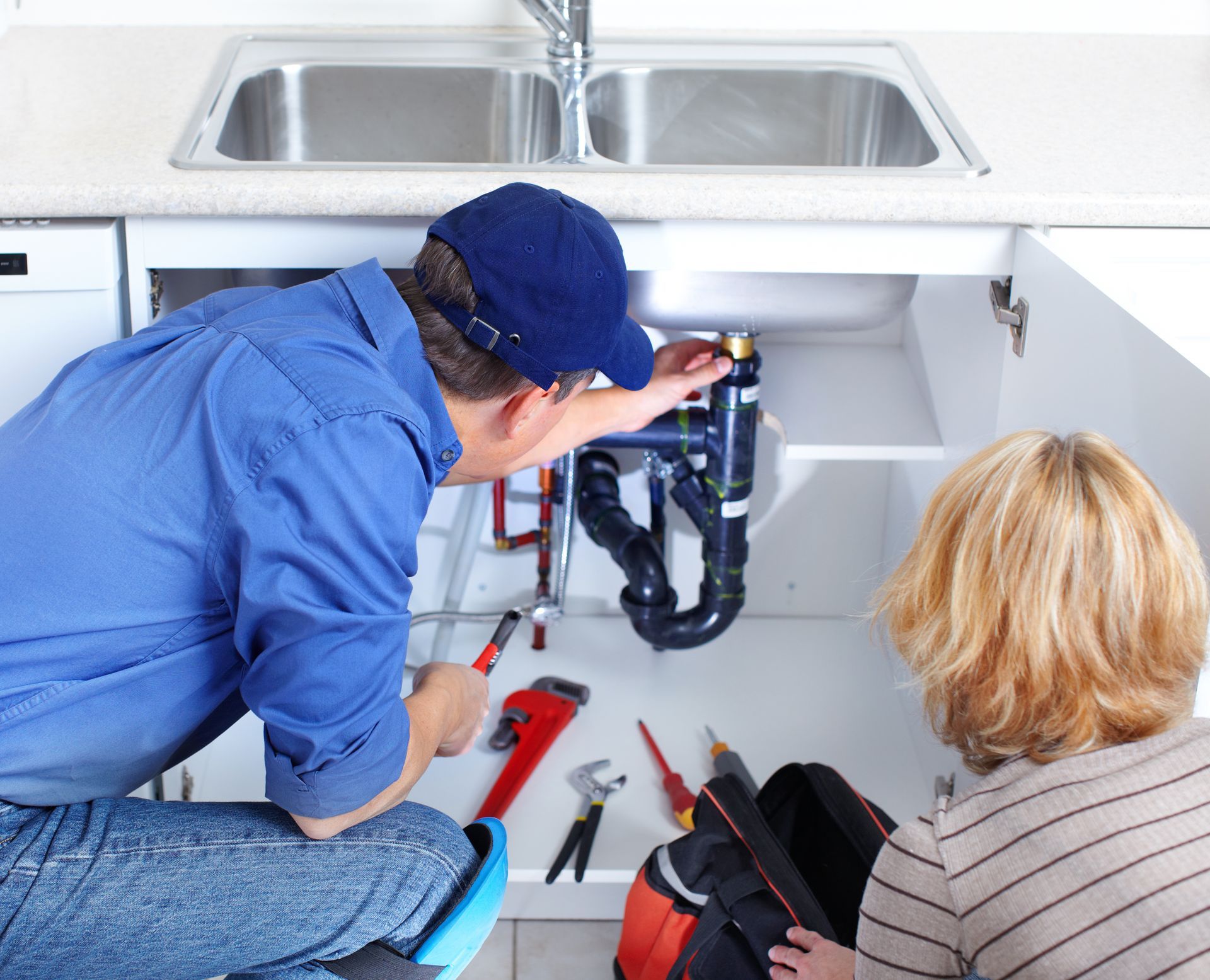 Plumber repairs pipes under a kitchen sink while a homeowner looks on.