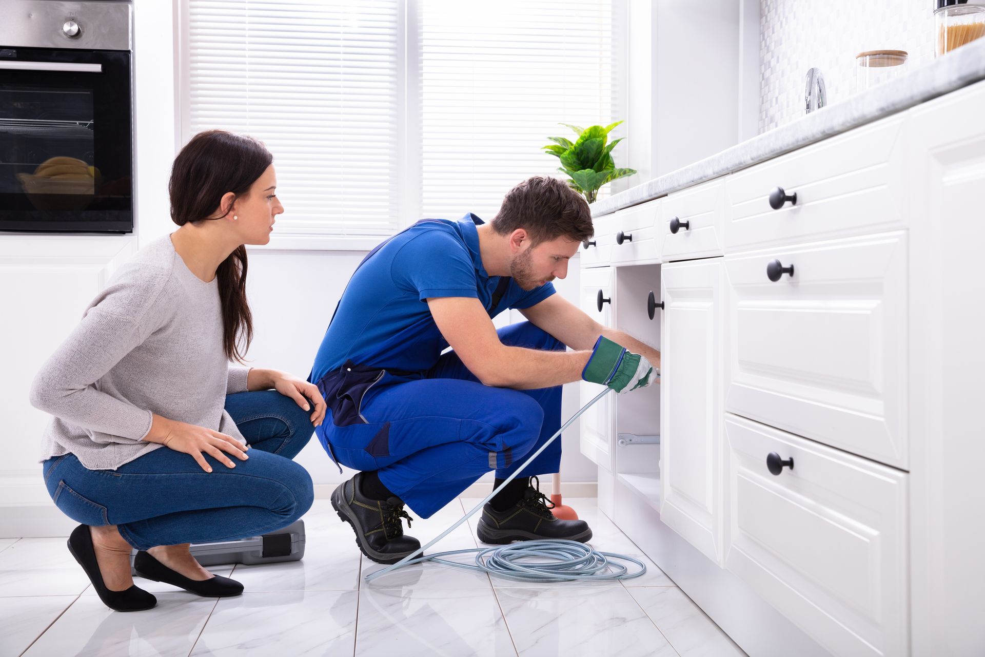 Plumber fixing kitchen sink pipes while homeowner observes in modern white cabinet kitchen.